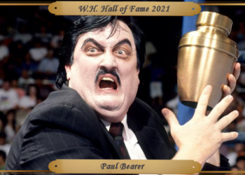 Black hair, dominant black eyebrows and bulging eyes against a powdered pale face, Paul Bearer scowls at the camera, clutching the golden urn of the Undertaker to the left of his body.