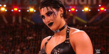 Rhea Ripley stares down her opponent wearing thick, black eye shadow and liner around the eyes, black lipstick and a black top with straps.