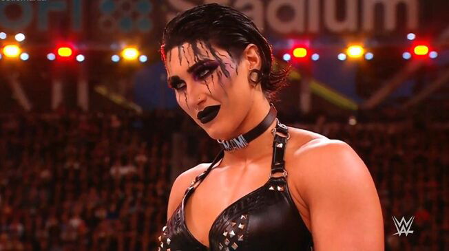 Rhea Ripley stares down her opponent from the wrestling ring, wearing thick, black eye shadow and liner around the eyes, black lipstick and a black top with straps.