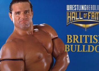 British Bulldog stands, posing with his left hand holding his right wrist, showing off his right bicep. He's wearing his trademark red, white and blue arm streamers.